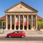 Kongress Palais Kassel mit klassischer Fassade und rotem Auto auf der Straße bei blauem Himmel.