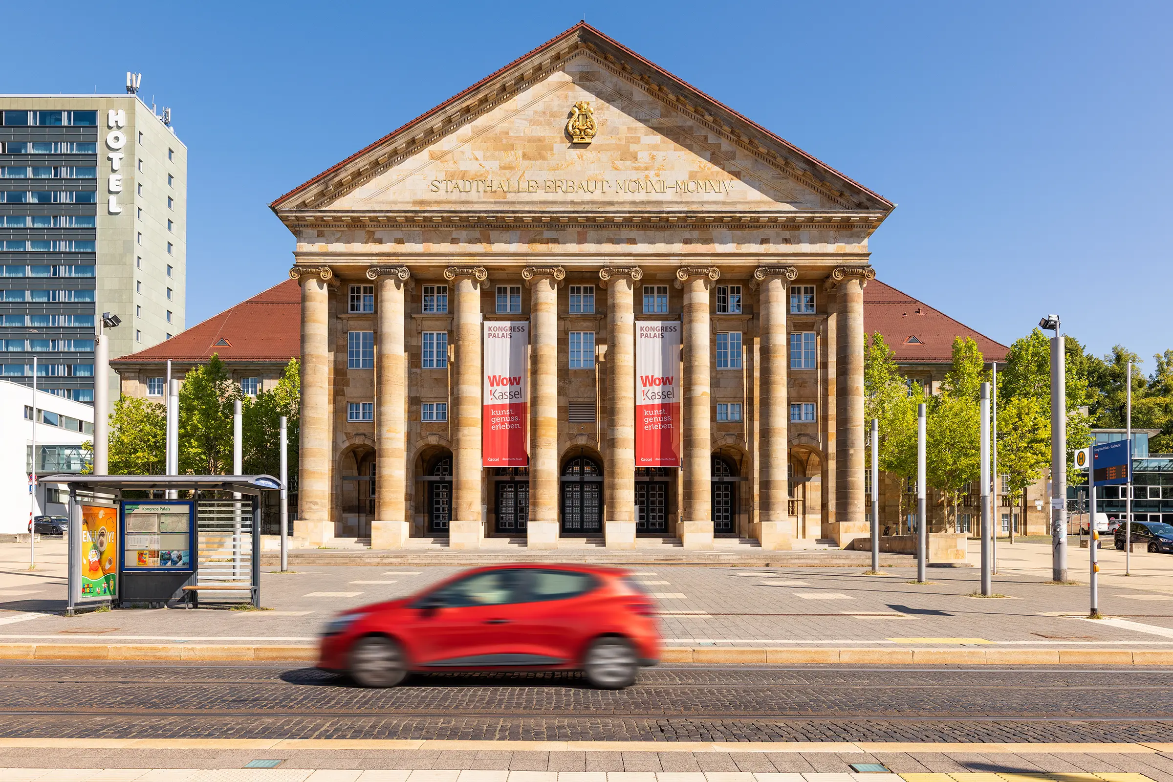 Kongress Palais Kassel mit klassischer Fassade und rotem Auto auf der Straße bei blauem Himmel.